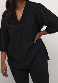 Blouse noire avec un col en V et une texture froncée au niveau du col. Dispose de manches trois-quarts et d'une coupe décontractée. Tissu lisse, design minimal.