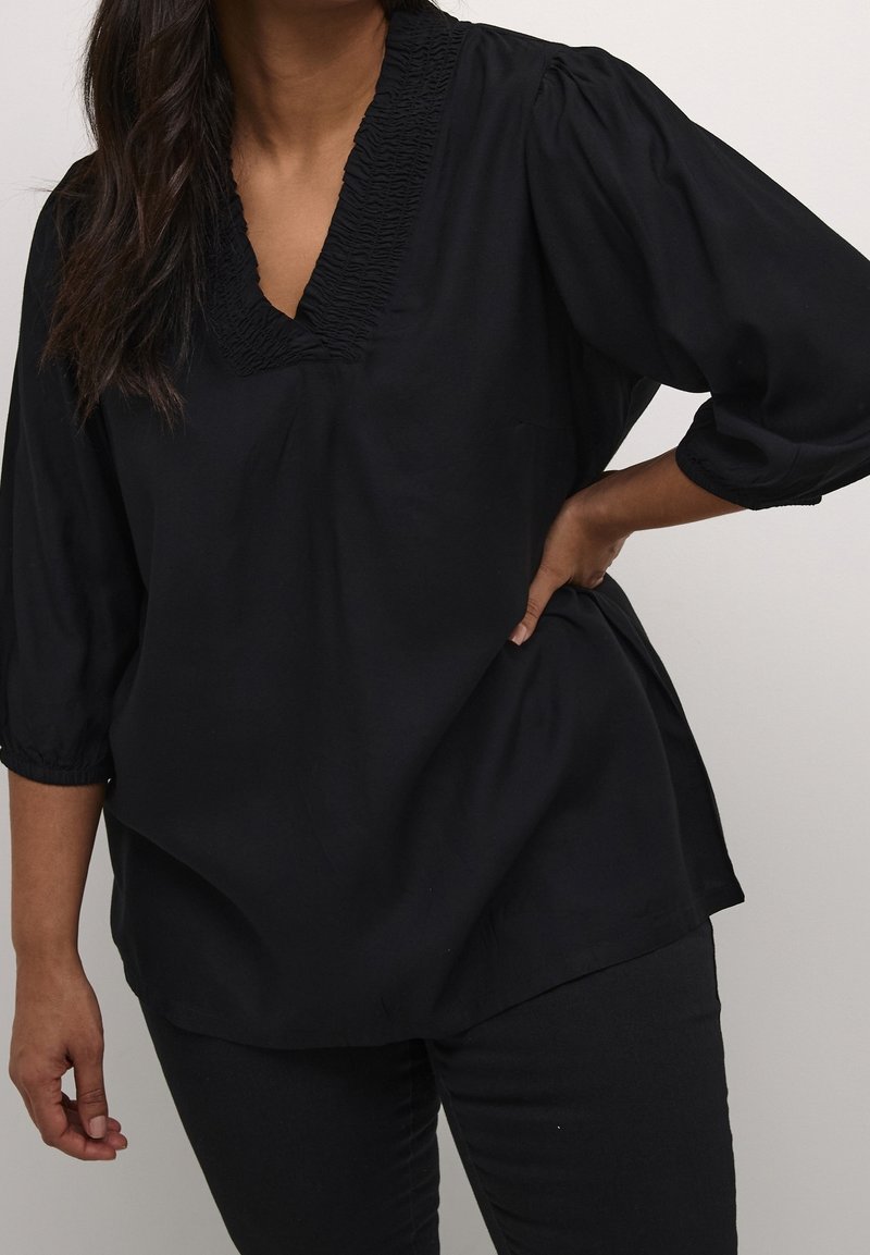 Blouse noire avec un col en V et une texture froncée au niveau du col. Dispose de manches trois-quarts et d'une coupe décontractée. Tissu lisse, design minimal.