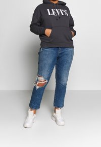 Personne portant un sweat à capuche Levi's noir, un jean bleu déchiré et des baskets blanches, debout les mains dans les poches du sweat, sur un fond uni.