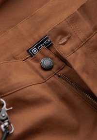 Bruine stoffen broek met een rits en een drukknoopsluiting. Bevat een zwart etiket met witte tekst en een metalen clip eraan bevestigd.