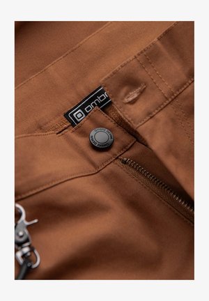 Pantalones de tela marrón con cierre de cremallera y botón a presión. Presentan una etiqueta negra con texto blanco y un clip de metal adjunto.