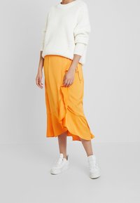 Maglione bianco lavorato a maglia con vestibilità over size abbinato a una gonna midi arricciata di un vivace arancione. Indossato con scarpe da ginnastica bianche su uno sfondo neutro.