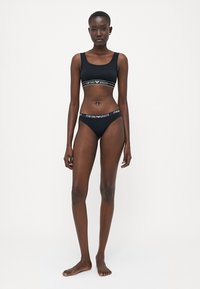 Fekete, két részből álló fehérnemű szett, amely egy logós pánttal ellátott cropped topból és hozzá illő bikinitról készült, mindkettő sima anyagból.