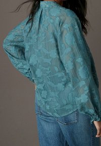 Una blusa leggera di colore teal con ricamo floreale, texture trasparente, maniche lunghe con polsini elasticizzati e scollo rotondo.
