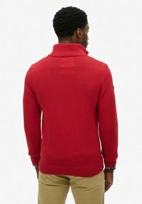 Suéter de punto rojo con un alto cuello acanalado, mangas largas y puños y dobladillo acanalados. Textura suave con un diseño ajustado.