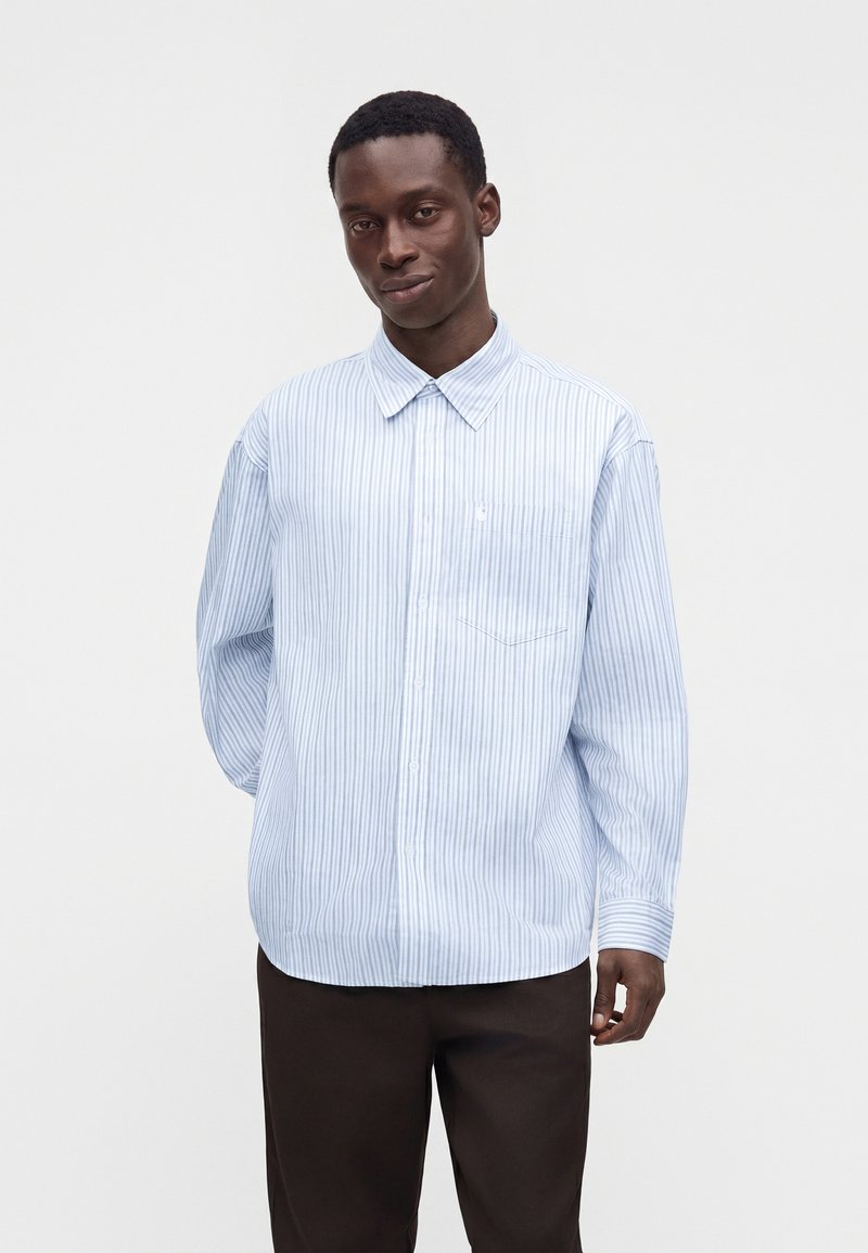 Uomo che indossa una camicia a righe verticali azzurro chiaro e bianca a manica lunga con colletto e pantaloni scuri, in piedi davanti a uno sfondo semplice.