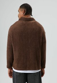 Pull-over en polaire marron avec une surface douce et texturée, col à revers et coupe décontractée. Le dos présente un ourlet droit et des manches longues.