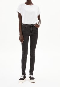 Personne portant un t-shirt blanc à manches courtes, un jean skinny noir taille haute et des baskets noires avec des semelles et des lacets blancs sur un fond blanc.