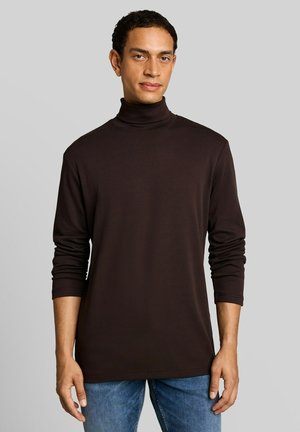 Mann trägt ein dunkelbraunes langärmeliges Rollkragenhemd und blaue Jeans und steht vor einem einfarbigen hellgrauen Hintergrund.