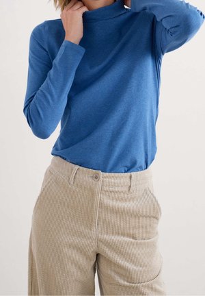 Persona con un top de cuello alto azul de manga larga metido dentro de unos pantalones de pana beige de talle alto con cierre de botón.