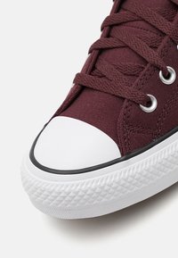 Mörkbruna kanvaslöparskor med vit gummikappa vid tårna och texturerad sula, med metallögla och matchande skosnören.