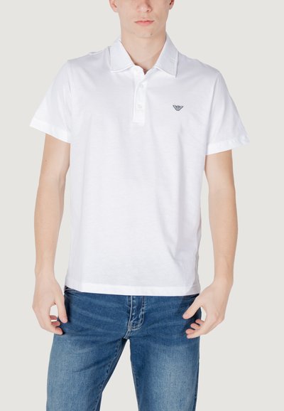 Polo blanc avec col, patte de boutonnage à deux boutons et logo noir brodé sur la poitrine gauche. Associé à un jean en denim bleu.