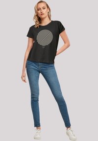 Schwarzes T-Shirt mit kurzen Ärmeln, das ein kreisförmiges geometrisches Design in Creme aufweist. Kombiniert mit engen blauen Jeans und weißen Sneakers.