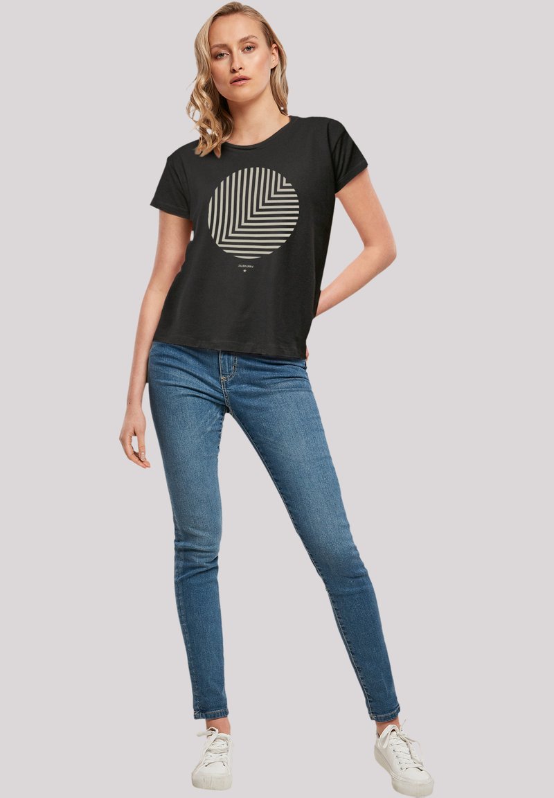 Schwarzes T-Shirt mit kurzen Ärmeln, das ein kreisförmiges geometrisches Design in Creme aufweist. Kombiniert mit engen blauen Jeans und weißen Sneakers.