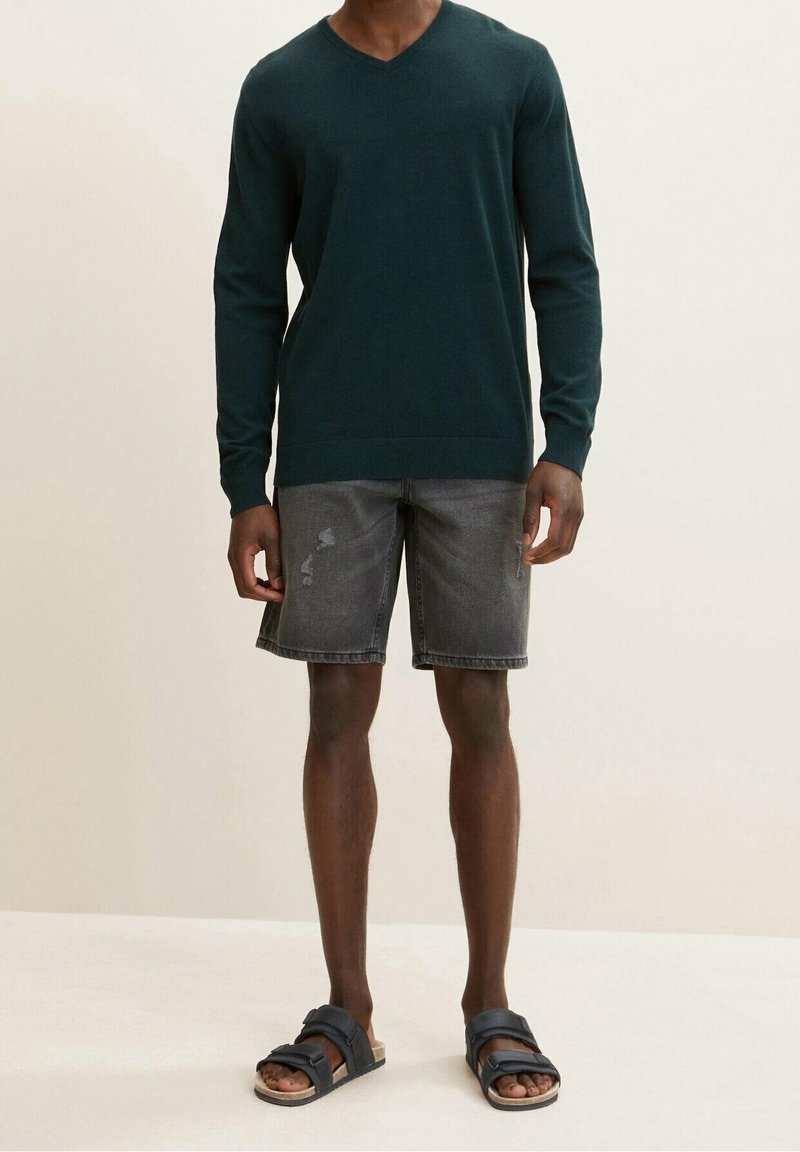 Dunkelgrüner V-Ausschnitt-Pullover mit langen Ärmeln, kombiniert mit ausgewaschenen Denim-Shorts und navy Sandalen mit Riemen-Design.