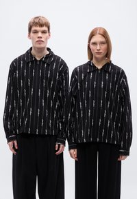 Μαύρα μπουφάν με φερμουάρ και κάθετα λευκά σχέδια κειμένου, με μεγάλους γιακάδες και άνετη εφαρμογή, φορεμένα πάνω από μαύρα φαρδιά παντελόνια.