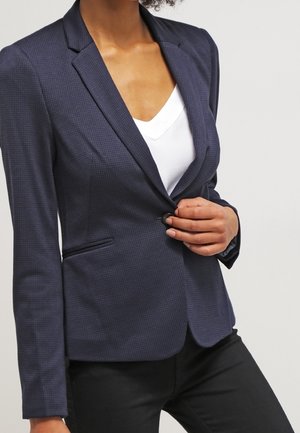 Femme fermant un blazer bleu foncé texturé sur une chemise blanche, portant un pantalon noir, sur un fond clair uni.