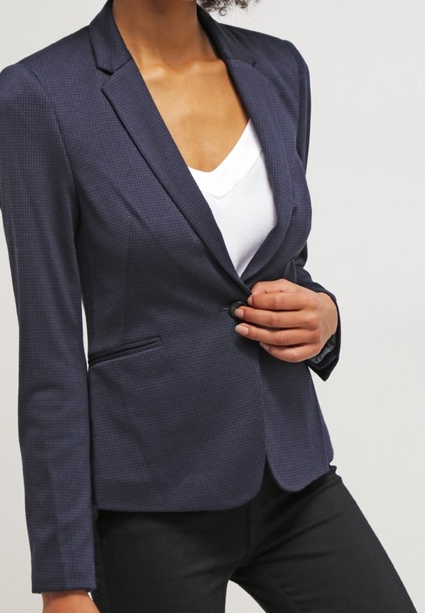 Femme fermant un blazer bleu foncé texturé sur une chemise blanche, portant un pantalon noir, sur un fond clair uni.