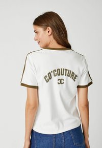 Balta trumpa rankovėmis marškinėliai su alyvuogių žalia apdaila ir kreiviu "CO'COUTURE" logotipu ant nugaros, pagaminti iš minkštos medvilnės medžiagos.