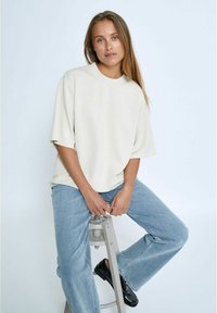 Camiseta oversized beige de mangas cortas, con cuello redondo y textura suave, combinada con jeans rectos de pierna azul clara y zapatos negros.