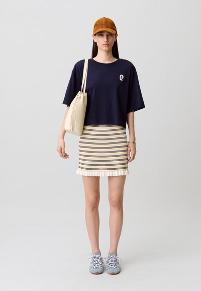Haut à manches courtes bleu marine avec logo, jupe rayée crème et bleu marine avec ourlet à volants, et baskets bleu clair. Un sac léger complète la tenue.