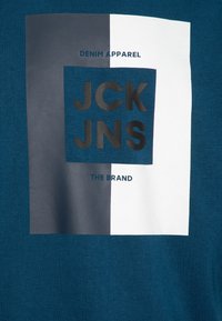 Camiseta turquesa con un gráfico rectangular con un fondo azul oscuro y blanco. El texto incluye "DENIM APPAREL" y "JCK JNS."