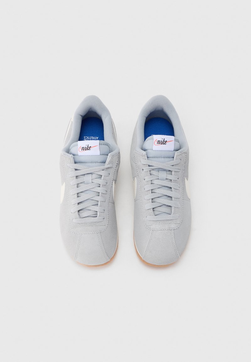 Light gray suede athletic shoes with flat laces, white logo accents, and a textured gum rubber sole. Interior features blue lining.