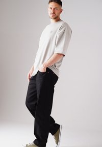 Hellgraues Übergrößen-T-Shirt mit kleinem Emblem, kombiniert mit locker sitzenden schwarzen Hosen und grünen Sneakers mit weißen Sohlen.