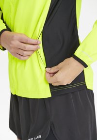 Hochsichtbare neon-gelbe Jacke mit schwarzem Seitenpanel, Reißverschluss und reflektierenden Akzenten, die von einer Hand von der Seite geöffnet wird.