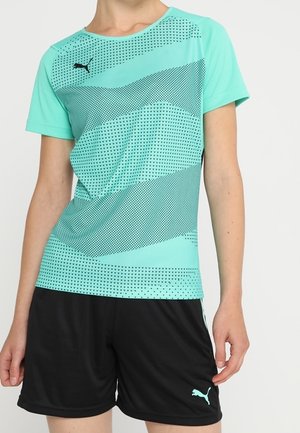 Person trägt ein türkisfarbenes Puma-Sport-T-Shirt mit schwarzen geometrischen Mustern und schwarze Puma-Shorts vor einem einfarbigen hellen Hintergrund.