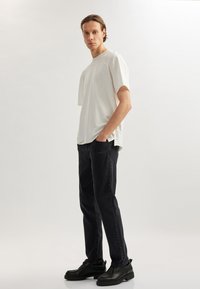 Camisa blanca de manga corta, ajuste relajado, combinada con jeans oscuros. El modelo lleva zapatos negros de acabado suave. Vestimenta simple y moderna.