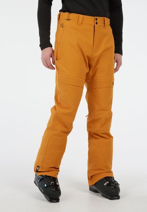 Pantalons de ski orange avec une texture lisse, comprenant deux poches latérales et un détail de fermeture éclair, associés à des bottes de ski noires.