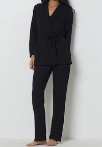 Il completo loungewear nero presenta una maglietta a involucro con cintura e maniche lunghe, abbinata a pantaloni dritti realizzati con lo stesso tessuto.