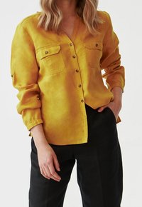 Camicia gialla senape con colletto a V, due tasche anteriori, maniche lunghe e tessuto a trama, abbinata a pantaloni neri.