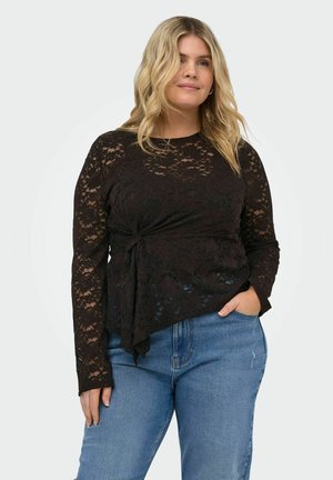 Blondynka ubrana w czarną koronkową bluzkę z długim rękawem z detalem węzła po boku i niebieskie dżinsy, stojąca z jedną ręką w kieszeni.