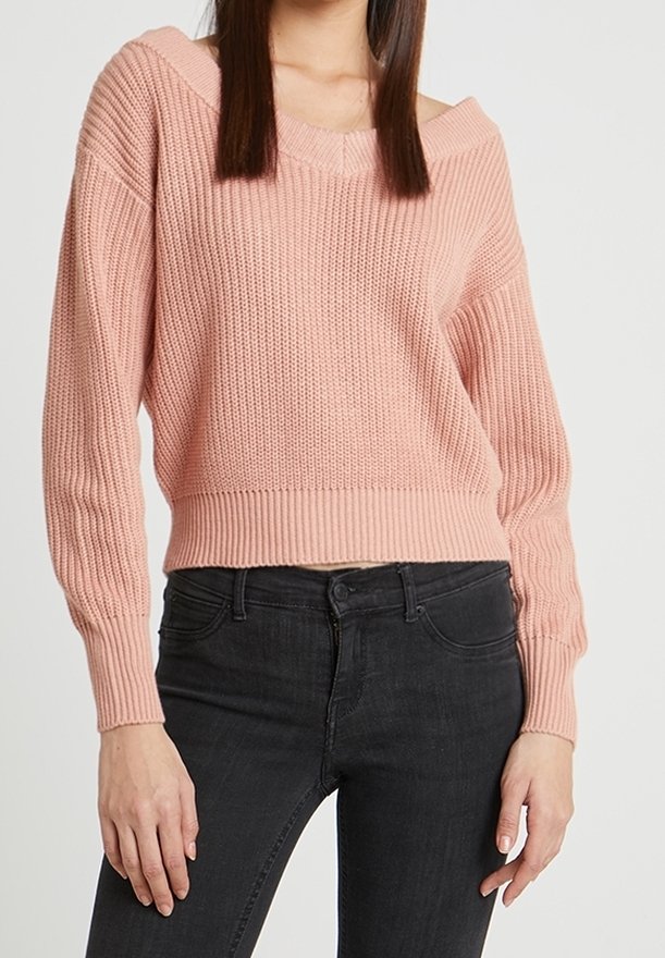 Femme portant un pull rose en tricot à manches longues associé à un jean noir taille haute sur un fond blanc.