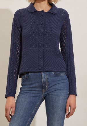 Kobieta ubrana w dopasowany granatowy kardigan z warkoczowym splotem i guzikami, zestawiony z niebieskimi dżinsami, na jednolitym tle.