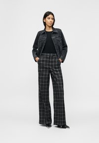 Γυναίκα που φοράει μαύρο τζιν μπουφάν, μαύρο τοπ, γκρι καρό παντελόνι με φαρδιά μπατζάκια και μαύρα παπούτσια με μυτερή μύτη, στέκεται με τα χέρια στις τσέπες.