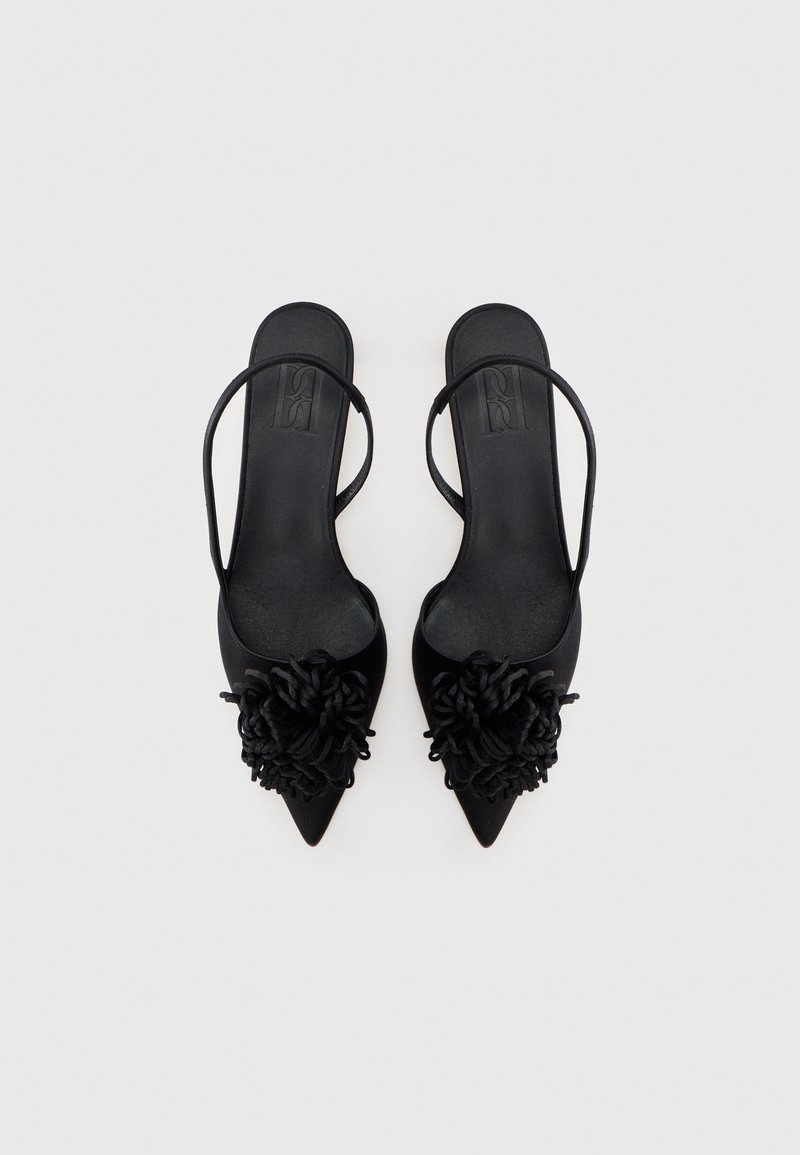 Sorte slingback-højhælede sko med spids snude, prydet med en stofblomst på vristen. Glat tekstur og minimalistiske designelementer.