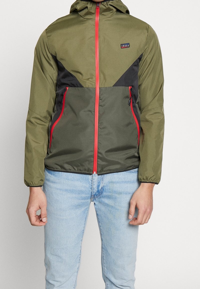 Lehká zelená bunda s černými akcenty a červenými zipy. Má kapuci, kapsy na zip a sportovní design. Viditelné logo na hrudi.