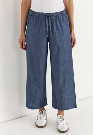 Kobieta nosząca luźne, szerokie spodnie z niebieskiego denimu z elastycznym pasem i białe trampki z brązowymi paskami.