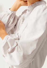 Blusa gris claro con mangas abullonadas voluminosas y cuello con volantes. Presenta cierre de botones en el cuello y una tela texturizada.
