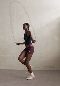 Woman in athletic wear jumping rope indoors against a plain wall, captured mid-jump with focused expression.