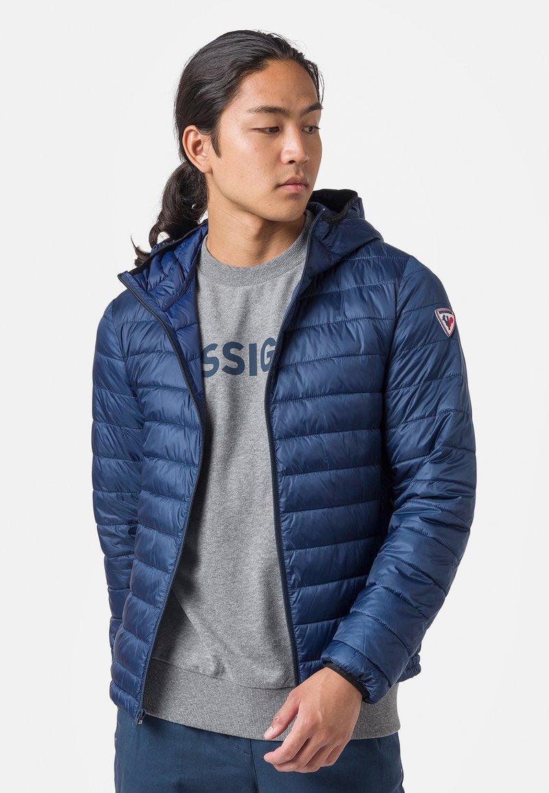 Giacca puffer blu navy con cappuccio, caratterizzata da cuciture orizzontali, chiusura frontale a zip e una piccola patch con logo sulla spalla sinistra.