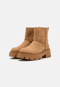 Botas de tobillo de ante color arena con suela de plataforma gruesa marrón, que presentan puntera redonda, paneles laterales elásticos y detalles de costura mínimos.