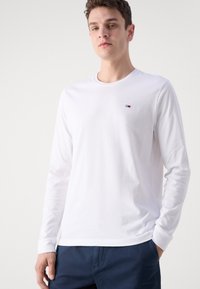 Camiseta de manga larga de algodón blanco con cuello redondo y un pequeño logotipo en el pecho. Textura suave y diseño ajustado.