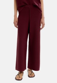 Burgundowe spodnie wide-leg z teksturowanym wykończeniem, z elastycznym pasem i sznurkiem do regulacji. Połączone z prostymi brązowymi sandałami.