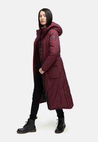 Bordeaux lang parka med hætte, lynlås og trykknapper, frontlommer og en glat tekstur, parret med sorte bukser og støvler.