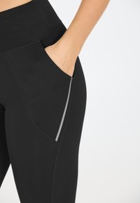 Gli leggings sportivi neri presentano una vita alta, una tasca laterale con rifiniture grigie e una trama di tessuto liscia e elastica.
