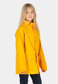 Jeune fille aux longs cheveux blonds portant une veste de pluie jaune vif avec poches à boutons et capuche, debout devant un fond uni.